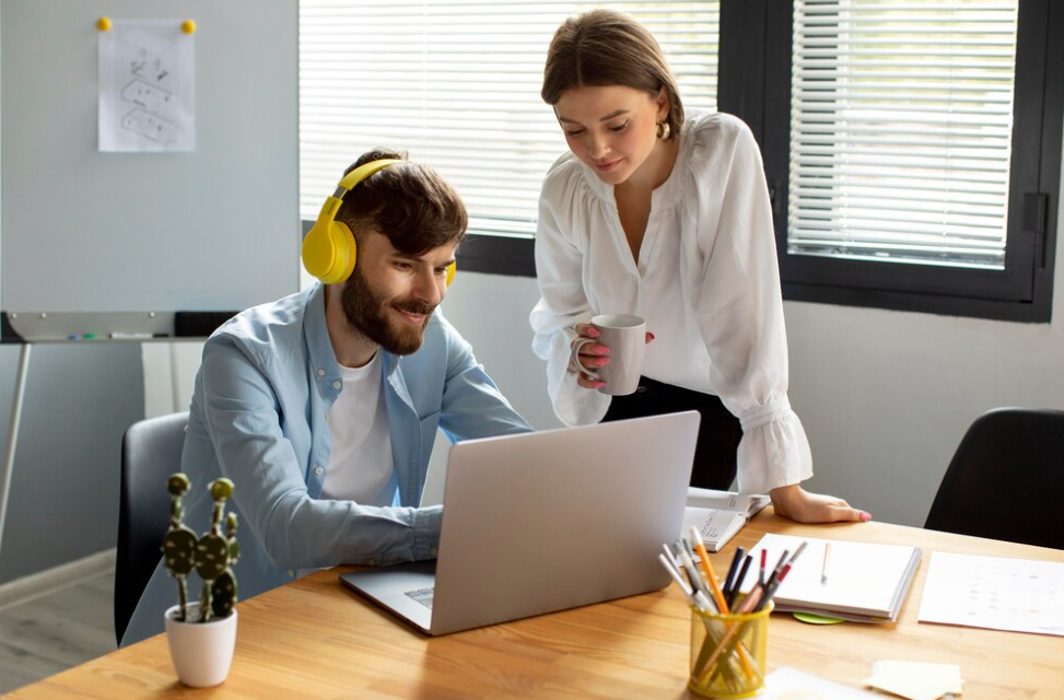 man with yellow headphones works using a laptop, a woman stands over him and looks at laptop