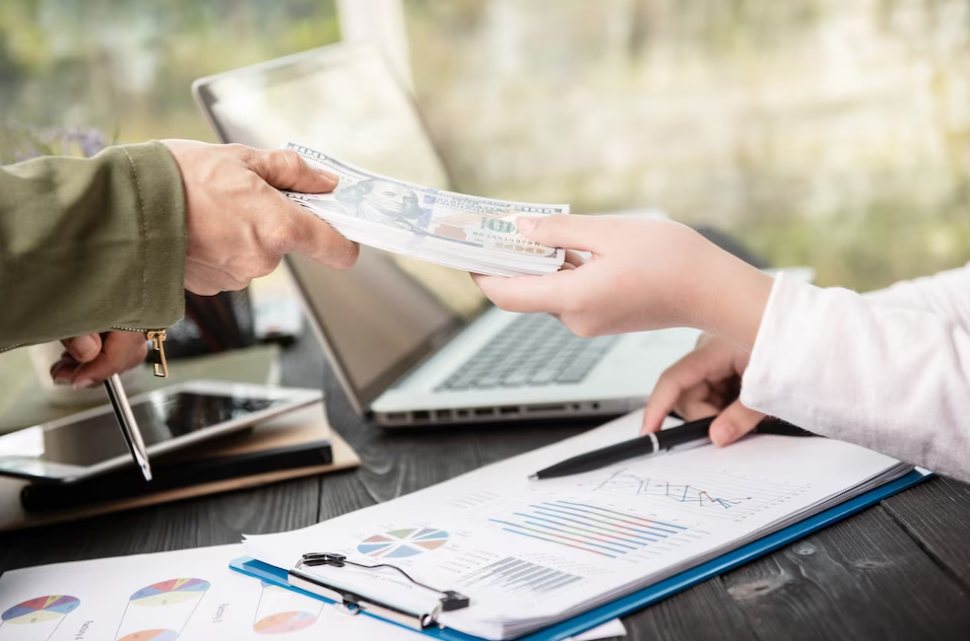 person’s hands passing the money to another, laptop and notes with graphs on the table